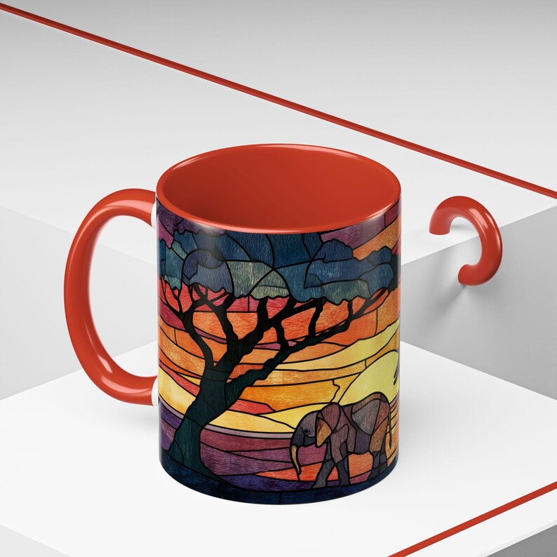 Puede incluir: Una taza de cer&aacute;mica con un vibrante dise&ntilde;o de puesta de sol africana. La taza presenta una colorida ilustraci&oacute;n de un &aacute;rbol y un elefante contra un fondo de puesta de sol. La taza tiene un asa y un interior rojos.