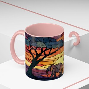 Puede incluir: Una taza de caf&eacute; de cer&aacute;mica con asa e interior rosas. El exterior presenta un dise&ntilde;o de vidriera de una escena de sabana africana con un &aacute;rbol, un elefante y una puesta de sol en tonos naranja, amarillo y azul.