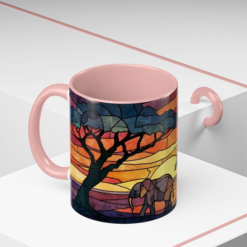 Puede incluir: Una taza de caf&eacute; de cer&aacute;mica con asa e interior rosas. El exterior presenta un dise&ntilde;o de vidriera de una escena de sabana africana con un &aacute;rbol, un elefante y una puesta de sol en tonos naranja, amarillo y azul.