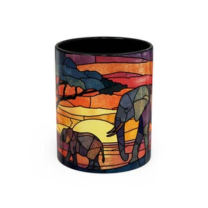 Puede incluir: Taza de cer&aacute;mica con un dise&ntilde;o de vidriera que representa dos elefantes en una sabana africana. La taza tiene un interior negro y un exterior colorido con tonos de naranja, amarillo, azul y morado.