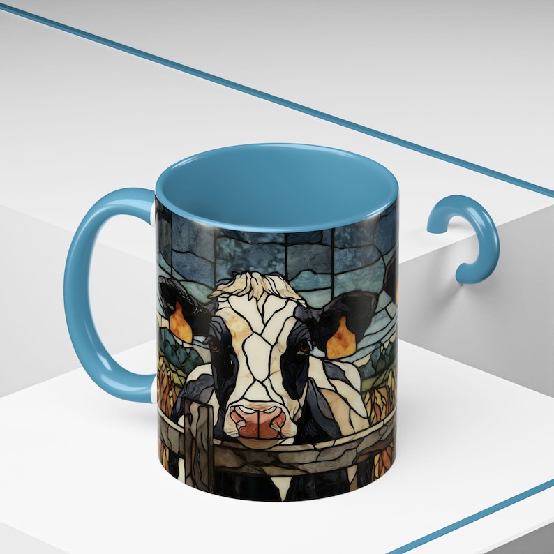 Puede incluir: Una taza de caf&eacute; de cer&aacute;mica con interior y asa azules. El exterior presenta una ilustraci&oacute;n estilo vidriera de una vaca asom&aacute;ndose por encima de una valla. La vaca es negra, blanca y marr&oacute;n, con nariz rosa y orejas naranjas.