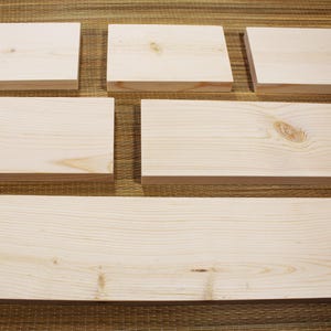 May include: A collection of unfinished, light-colored wooden planks and blocks. The pieces vary in size, with several rectangular blocks and a long, wide plank. The wood grain is visible, and the pieces are arranged on a textured surface.