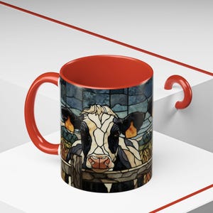 Puede incluir: Una taza de caf&eacute; de cer&aacute;mica con interior y asa rojos. El exterior presenta una imagen de estilo vidriera de una vaca mirando por encima de una valla. La vaca es negra, blanca y marr&oacute;n, con una nariz rosa. El fondo es azul y verde.