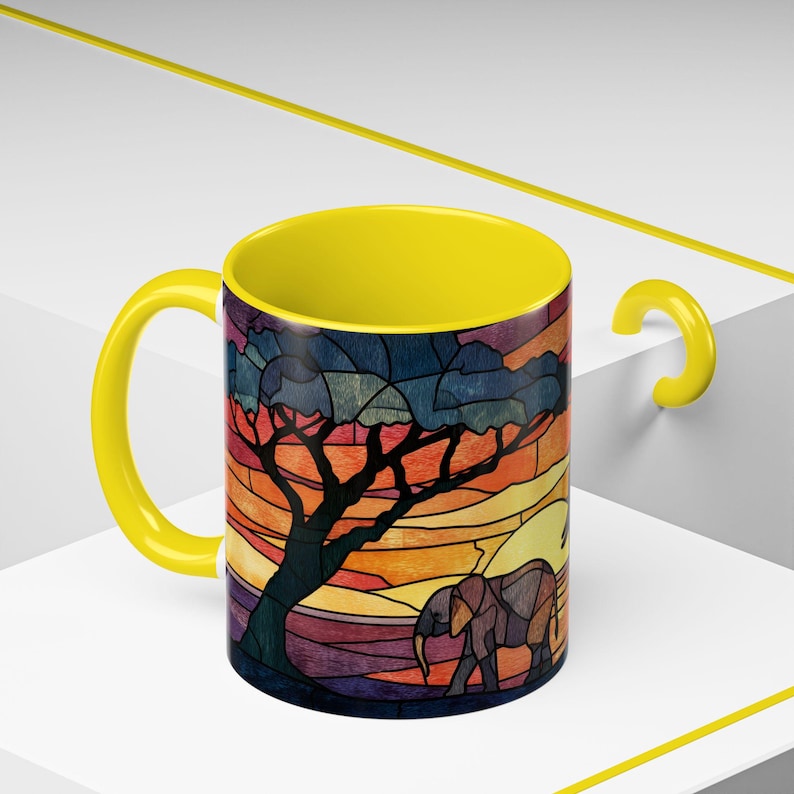 Puede incluir: Una taza de caf&eacute; con asa amarilla, con un dise&ntilde;o de vidriera que representa un &aacute;rbol y un elefante sobre un fondo de puesta de sol. El interior de la taza es de color amarillo brillante, complementando el asa y los colores c&aacute;lidos del dise&ntilde;o.