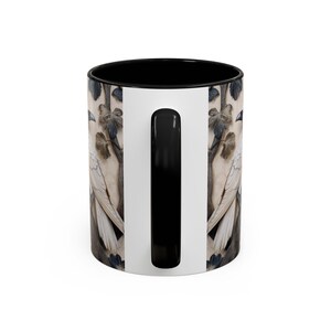 May include: A ceramic coffee mug with a black interior and handle. The exterior features a detailed illustration of a white bird perched on a branch with leaves. The background is a neutral beige color.