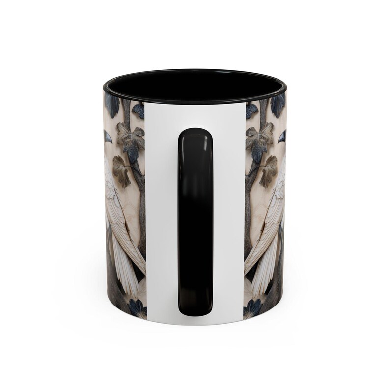 May include: A ceramic coffee mug with a black interior and handle. The exterior features a detailed illustration of a white bird perched on a branch with leaves. The background is a neutral beige color.