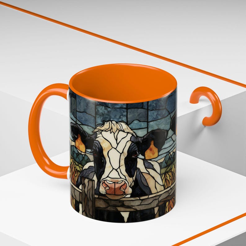 Puede incluir: Taza de caf&eacute; con asa e interior naranja, decorada con un dise&ntilde;o de vaca estilo vidriera. La vaca es blanca y negra con una nariz rosa, mirando por encima de una valla de madera. El fondo es un patr&oacute;n de vidriera azul y verde.