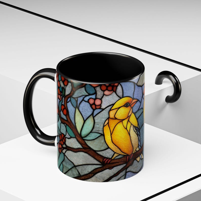Puede incluir: Taza de cer&aacute;mica negra con un dise&ntilde;o de vidriera de un p&aacute;jaro amarillo posado en una rama con bayas rojas y hojas verdes. La taza tiene un interior y un asa negros.