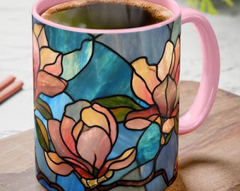 May include: A ceramic coffee mug with a pink handle and rim, filled with coffee. The mug features a stained-glass design of pink, peach, and yellow magnolia flowers against a blue background. The mug sits on a wooden surface.