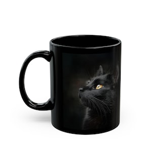 Pu&ograve; includere: Tazza in ceramica nera con l'immagine di un gatto nero con occhi giallo brillante. Il gatto guarda di lato su uno sfondo scuro. La tazza ha un manico curvo.