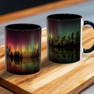 May include: Two ceramic mugs with a scenic design. The design features a colorful aurora borealis over a lake with silhouetted trees. One mug has a black handle and rim, the other has a black rim.