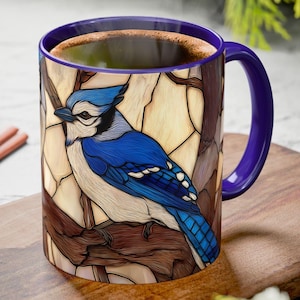 May include: A ceramic mug with a blue jay design. The mug features a vibrant blue jay perched on a branch, set against a stained-glass style background. The mug has a purple handle and interior, and is filled with a dark liquid.