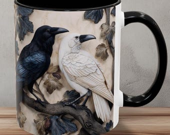 May include: A ceramic mug with a black handle and interior. The mug features a detailed relief of two ravens, one black and one white, perched on a branch with leaves. The background is a neutral beige.