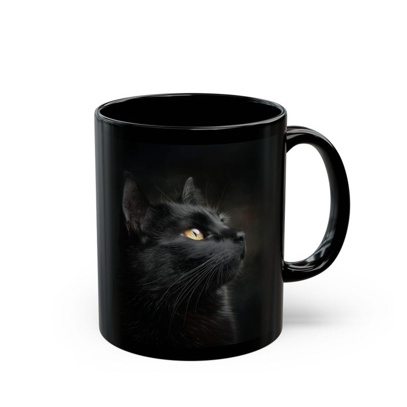 Pu&ograve; includere: Tazza in ceramica nera con un'immagine dettagliata di un gatto nero con occhi gialli. Il gatto guarda verso l'alto, su uno sfondo scuro. La tazza ha un manico curvo e una finitura lucida, ideale per caff&egrave; o t&egrave;.