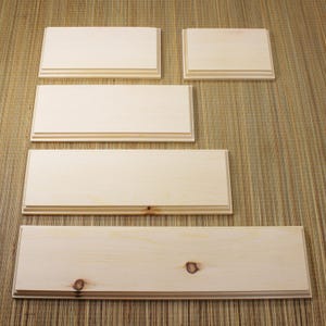 May include: A collection of unfinished wooden planks and panels, varying in size, arranged on a textured, light brown surface. The wood has a natural, light color with visible grain and knots, suggesting a DIY or craft project.