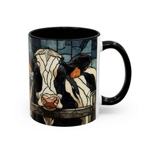 Puede incluir: Una taza de cer&aacute;mica blanca con interior y asa negros. La taza presenta una colorida ilustraci&oacute;n de estilo vidriera del rostro de una vaca. La vaca es blanca y negra con una nariz rosada y est&aacute; detr&aacute;s de una valla de madera.