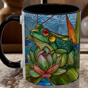 May include: A ceramic coffee mug with a black handle and interior. The mug features a stained-glass style design of a green frog with red and yellow eyes, sitting on a lily pad. The background is blue with green and orange accents.