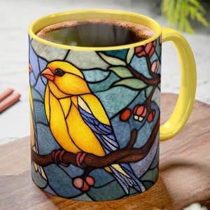 May include: A yellow ceramic mug with a stained-glass design featuring a yellow bird perched on a branch with red berries. The mug has a yellow handle and rim, and is filled with a dark liquid.