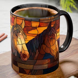 May include: A black-handled coffee mug featuring a stained-glass design of two horses. The horses are brown and tan, set against a warm orange and yellow background. The mug is filled with a dark liquid.