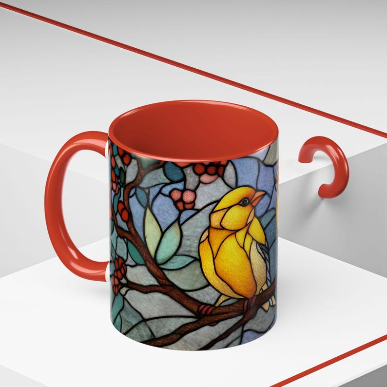Puede incluir: Taza de cer&aacute;mica con un dise&ntilde;o de vidriera que presenta un p&aacute;jaro amarillo posado en una rama con bayas rojas. El interior y el asa son rojos. La taza est&aacute; sobre un fondo blanco con detalles rojos.