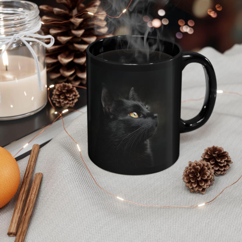 Pu&ograve; includere: Tazza in ceramica nera con un ritratto di un gatto nero con occhi gialli. La tazza ha un manico curvo ed &egrave; riempita con un liquido scuro, con vapore che sale dalla parte superiore. La tazza &egrave; su una superficie grigio chiaro.