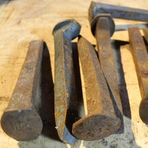 May include: A collection of rusty metal railroad spikes, some with square heads and some with round heads. The spikes are arranged on a wooden surface.