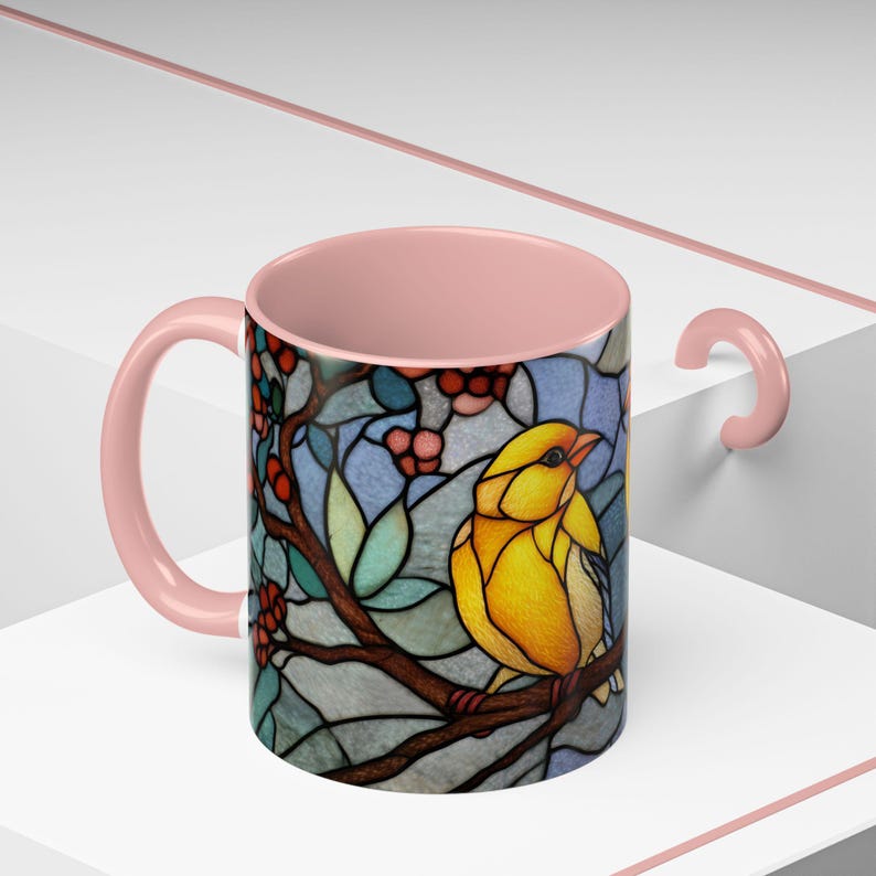 Puede incluir: Una taza de cer&aacute;mica con asa e interior rosas. El exterior presenta un dise&ntilde;o de vidriera de un p&aacute;jaro amarillo posado en una rama con bayas rojas y hojas verdes sobre un fondo azul.