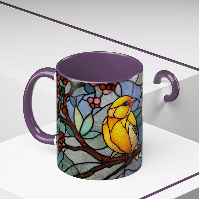 Puede incluir: Una taza de cer&aacute;mica con un dise&ntilde;o de vidriera que presenta un p&aacute;jaro amarillo posado en una rama con bayas rojas. El interior y el asa son de color p&uacute;rpura oscuro. La taza est&aacute; sobre un fondo blanco.
