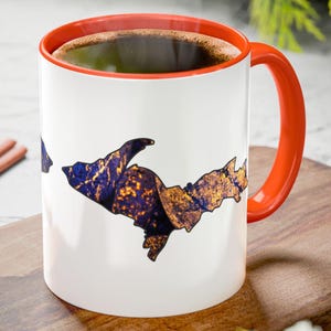 May include: White ceramic coffee mug with an orange handle and interior. The mug features a graphic of a landmass in shades of blue, gold, and purple. The mug is filled with coffee.
