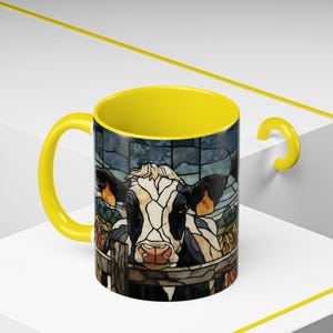 Puede incluir: Una taza de caf&eacute; de cer&aacute;mica con asa e interior amarillos. El exterior presenta una ilustraci&oacute;n de estilo vidriera de una vaca mirando por encima de una valla. La vaca tiene marcas blancas y negras y una nariz rosada. El fondo es un paisaje azul y verde.