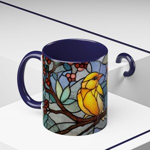 Puede incluir: Una taza de cer&aacute;mica con interior, asa y borde azul marino. El exterior presenta un dise&ntilde;o de vidriera de un p&aacute;jaro amarillo posado en una rama con bayas rojas y hojas verdes. El fondo es una mezcla de azul y blanco.