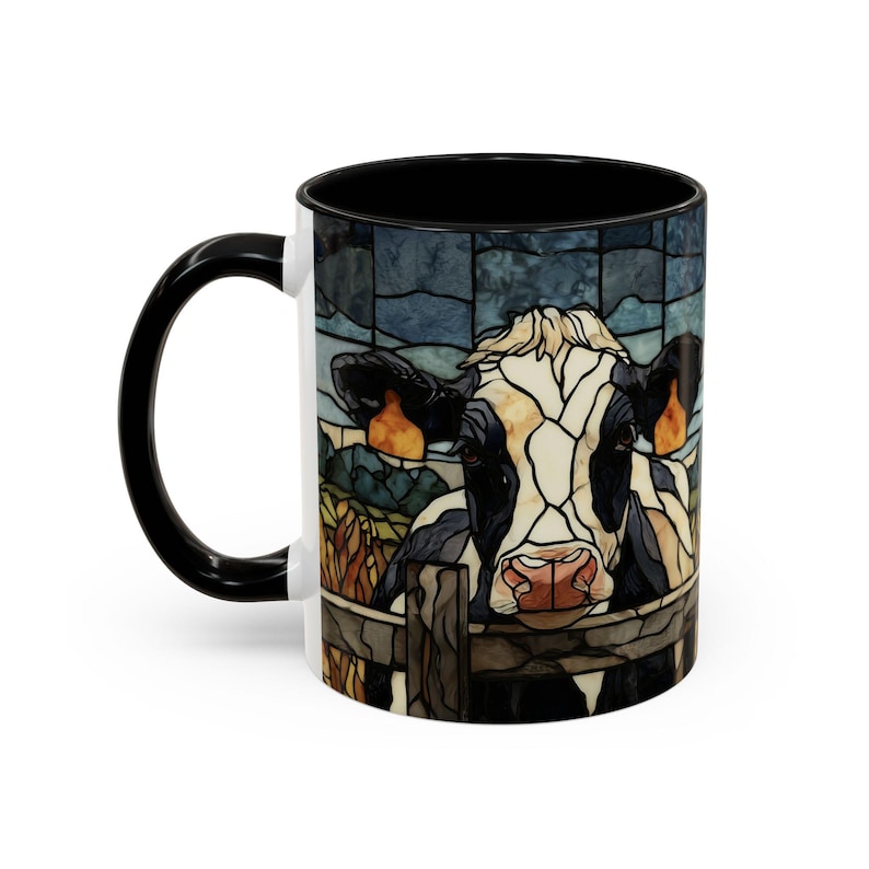 Puede incluir: Una taza de caf&eacute; de cer&aacute;mica con asa e interior negros. La taza presenta una ilustraci&oacute;n de estilo vidriera de una vaca que mira por encima de una valla de madera. La vaca es blanca y negra con nariz rosa y orejas naranjas.