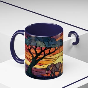 Puede incluir: Una taza de cer&aacute;mica con asa e interior azul oscuro. El exterior presenta un colorido dise&ntilde;o de vidriera de una escena de sabana africana con un &aacute;rbol, una puesta de sol y un elefante. La taza est&aacute; sobre un fondo blanco.