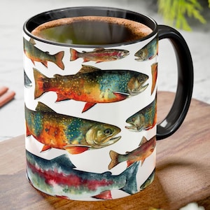 May include: A white ceramic coffee mug with a black handle and rim. The mug is decorated with watercolor illustrations of various colorful fish. The mug is filled with a dark brown liquid, possibly coffee or tea.