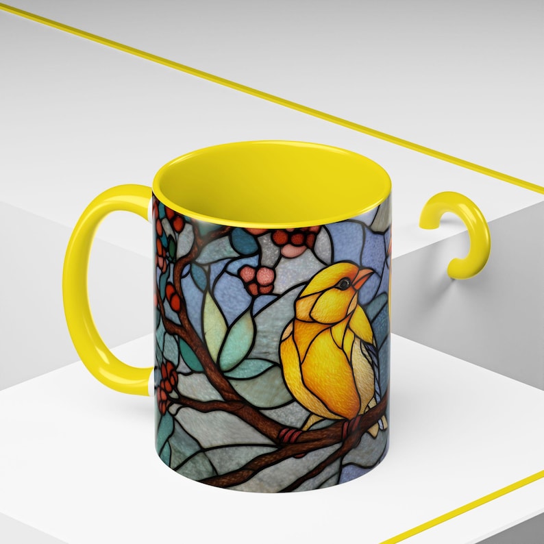 Puede incluir: Una taza de cer&aacute;mica amarilla con un dise&ntilde;o de vidriera que presenta un p&aacute;jaro amarillo posado en una rama con bayas rojas y hojas verdes. El interior y el asa de la taza son de color amarillo brillante.