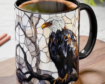 May include: A ceramic coffee mug with a black handle and rim. The mug features a stained-glass style design of a bald eagle perched on a branch. The eagle has white feathers on its head and a yellow beak. The mug is filled with coffee.
