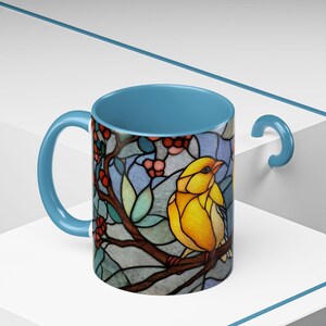 Puede incluir: Una taza de cer&aacute;mica con interior y asa azul claro. El exterior presenta un dise&ntilde;o de vidriera de un p&aacute;jaro amarillo posado en una rama con bayas rojas y hojas verdes. La taza est&aacute; sobre un fondo blanco.