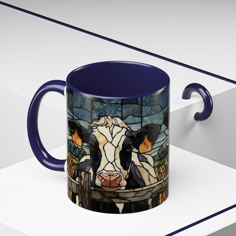Puede incluir: Una taza de caf&eacute; de cer&aacute;mica con interior y asa azul marino. El exterior presenta una ilustraci&oacute;n estilo vidriera de una vaca asom&aacute;ndose por encima de una valla. La vaca es negra, blanca y marr&oacute;n claro, con una nariz rosada. El fondo es un dise&ntilde;o de vidriera azul.