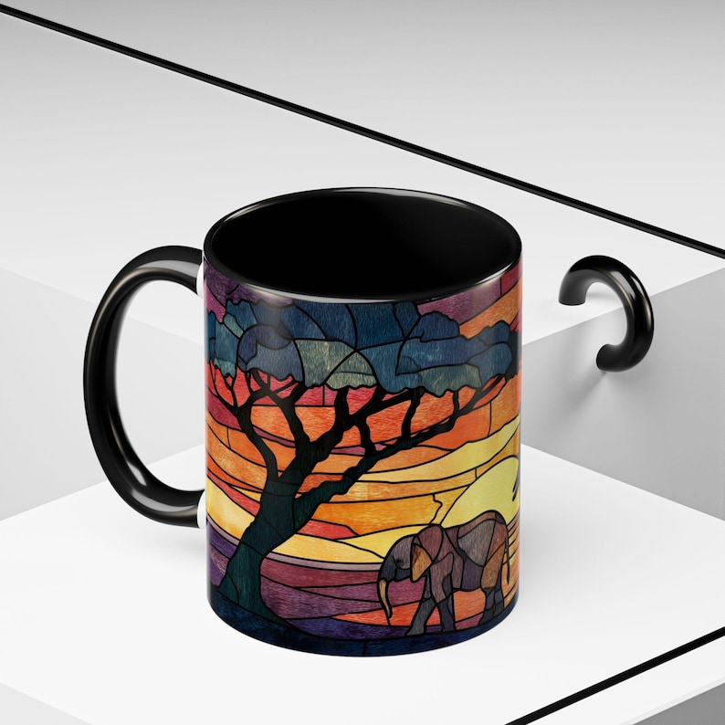 Puede incluir: Taza de cer&aacute;mica negra con un dise&ntilde;o de vidriera que representa una sabana africana. El dise&ntilde;o incluye un &aacute;rbol, un elefante y una puesta de sol en tonos naranja, amarillo y morado. La taza tiene un asa y un interior negros.