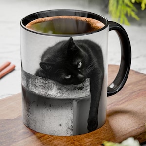May include: A black and white ceramic mug featuring a black cat resting on a concrete ledge. The mug has a black handle and rim, and is filled with a dark liquid. The cat's eyes are visible, and the image is printed on the mug's surface.