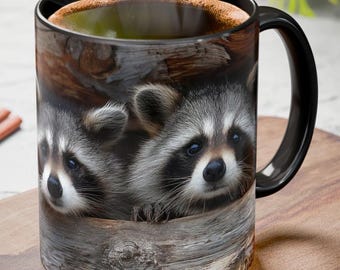 May include: Black coffee mug featuring a detailed image of two raccoons peering out from a wooden log. The mug has a black handle and rim, and is filled with a hot beverage. The mug is sitting on a wooden surface.