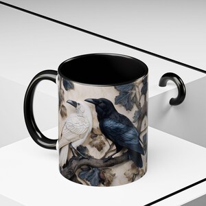 May include: A black and white ceramic mug featuring a detailed relief of two ravens perched on a branch. One raven is white, the other is black. The mug has a black interior and handle, with a cream-colored background.