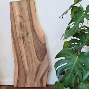 May include: A large, natural wood slab with a live edge and a unique grain pattern. The wood is a light brown color with darker brown streaks. The slab is sitting on a wooden floor.