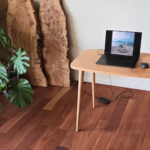 May include: A light wood table with a laptop computer, a wireless mouse, and a power adapter on the surface. The table has four legs and is sitting on a hardwood floor. A potted plant is in the foreground.