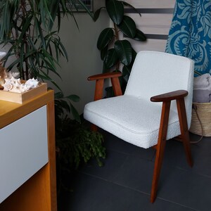 May include: A white armchair with wooden legs and arms. The chair is in a room with a plant and a blue patterned blanket.