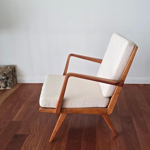 May include: A wooden armchair featuring a light-toned, textured fabric seat and backrest cushion. The chair has a natural wood frame with angled armrests and tapered legs, showcasing a clear wood grain finish throughout the structure.