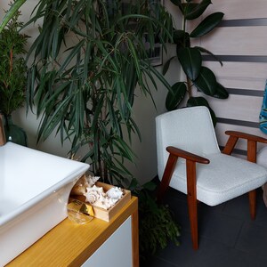 May include: A white armchair with wooden arms and legs sits in a bathroom with a white porcelain sink and a wooden countertop.  The bathroom has a variety of plants, including a large green plant in the corner and a smaller plant on the countertop.  There is a blue and white patterned towel hanging on the wall.