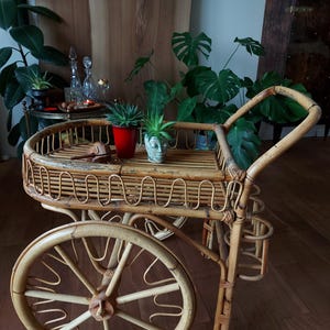 May include: A vintage bamboo serving cart with large spoked wheels. The cart has a rectangular top with a curved edge and a lower shelf. It holds potted plants and decorative items. The cart is made of light brown bamboo.