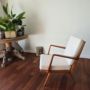 以下が含まれることがあります： 明るい色のクッションと木製フレームを備えたミッドセンチュリーモダンアームチェア。椅子は角度の付いた脚とアームレストを備えています。部屋には、植物のある丸い木製のテーブルと濃い色の木の床があります。