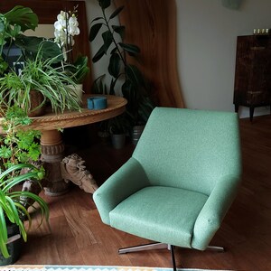 May include: A green swivel chair with a modern design sits on a red and white patterned rug in a living room. The chair is in front of a wooden table with plants and a wooden wall panel.